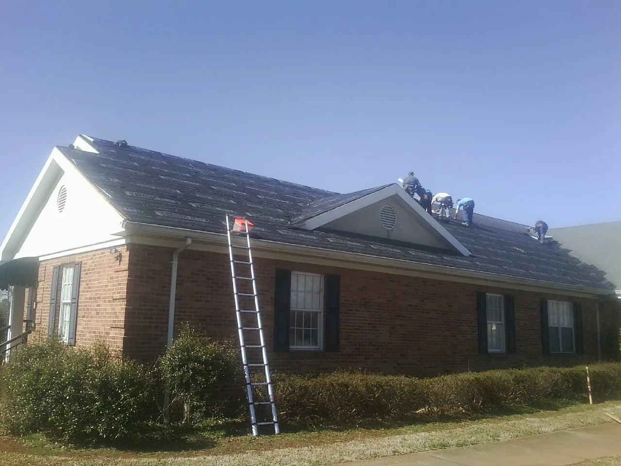 Crew tearing shingles off a brick ranch roof with a ladder set — Metal Roof Repair work by our crew in Milburn