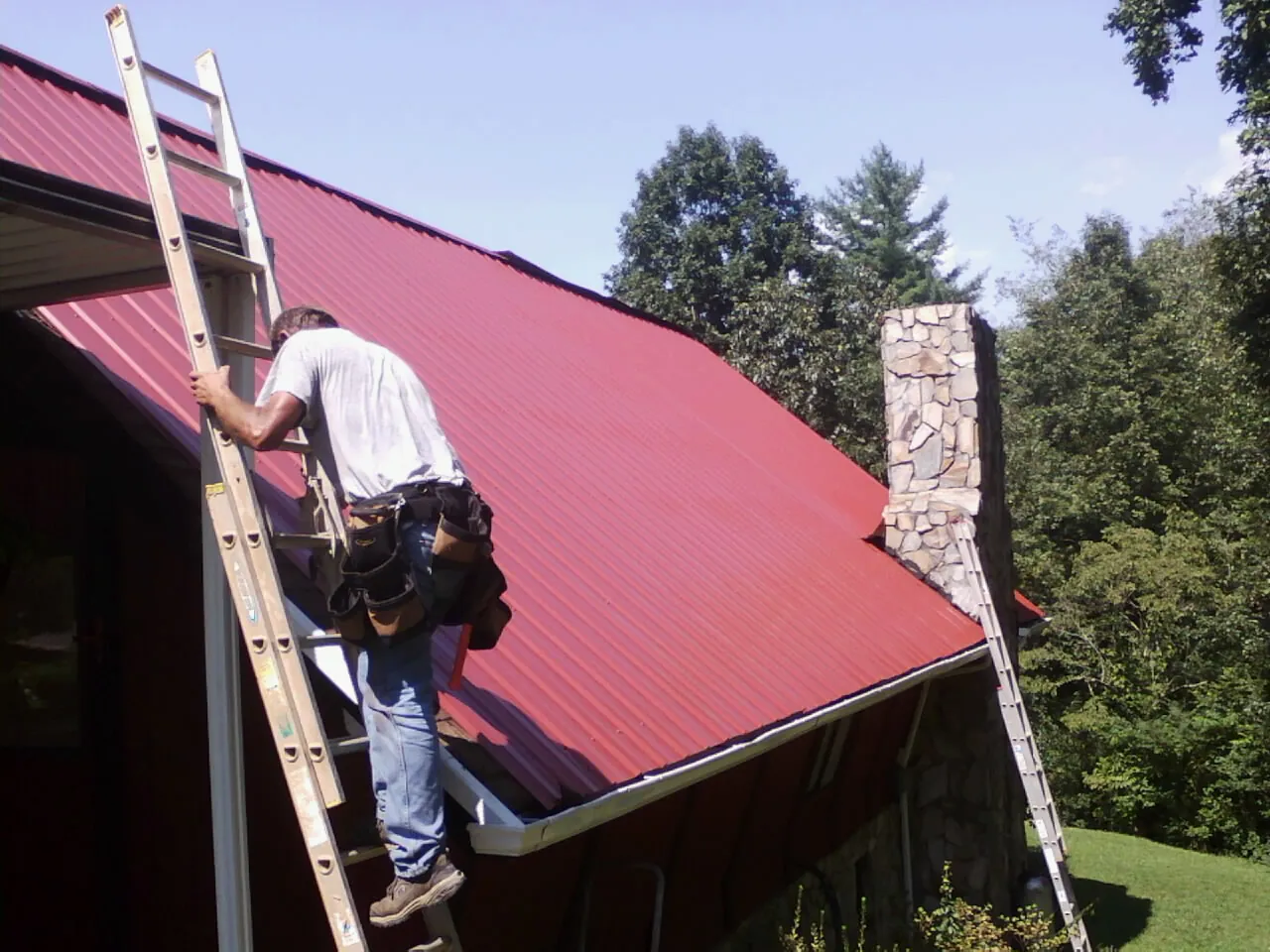 Roofer on a ladder beside a red metal roof and stone chimney — Soffit & Fascia Repair by Roofing Brothers in Milburn